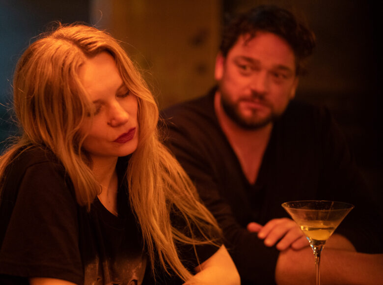 W pomieszczeniu kobieta i mężczyzna siedzą przy stole. Na stole stoi jeden kieliszek z napojem.