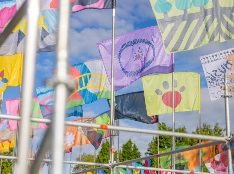 Kilkanaście kolorowych chorągwi przyczepionych do rusztowania. W tle niebo i drzewa.