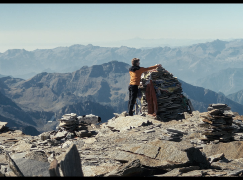Widok na skaliste szczyty gór. Na bliższym planie na jednym ze szczytów stoi mężczyzna oparty o stertę kamiennych płyt.