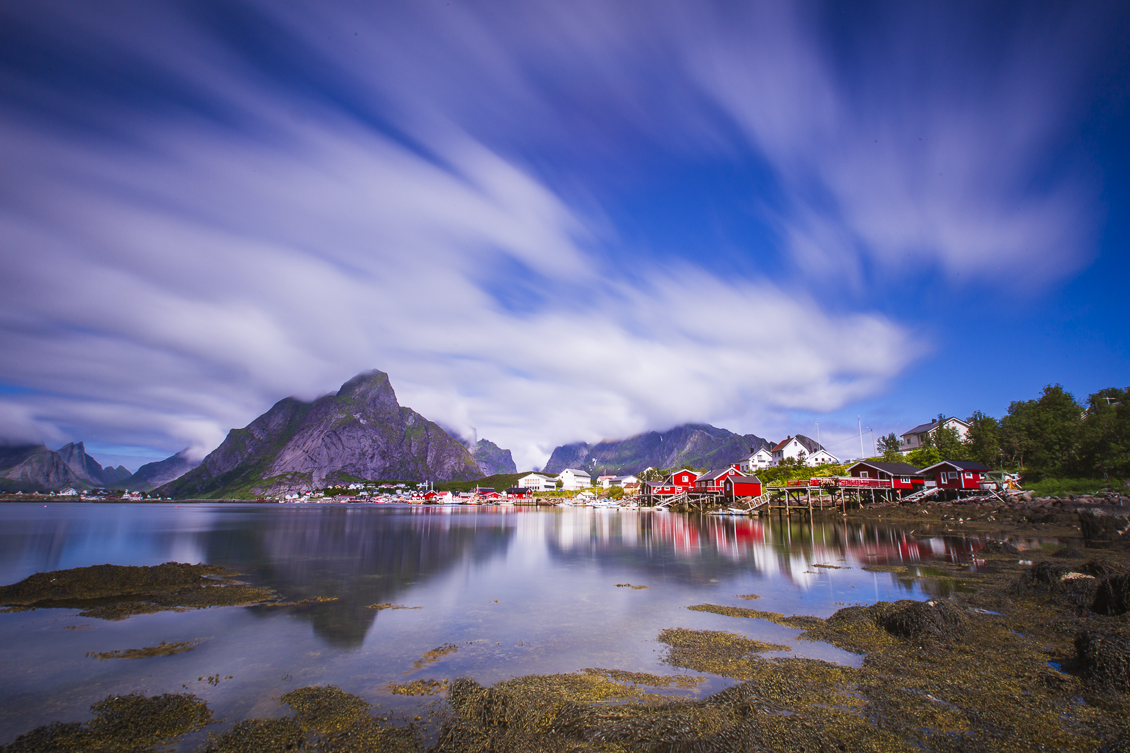 Kolorowa fotografia przedstawia widok fiordu otoczonego wysokimi górami. Typowy północny krajobraz wody, niebieskiego nieba i&nbsp;gór, kontrastuje z&nbsp;pomalowanymi na&nbsp;czerwono drewnianymi zabudowaniami, również bardzo charakterystycznymi dla&nbsp;krajobrazu północy Europy.