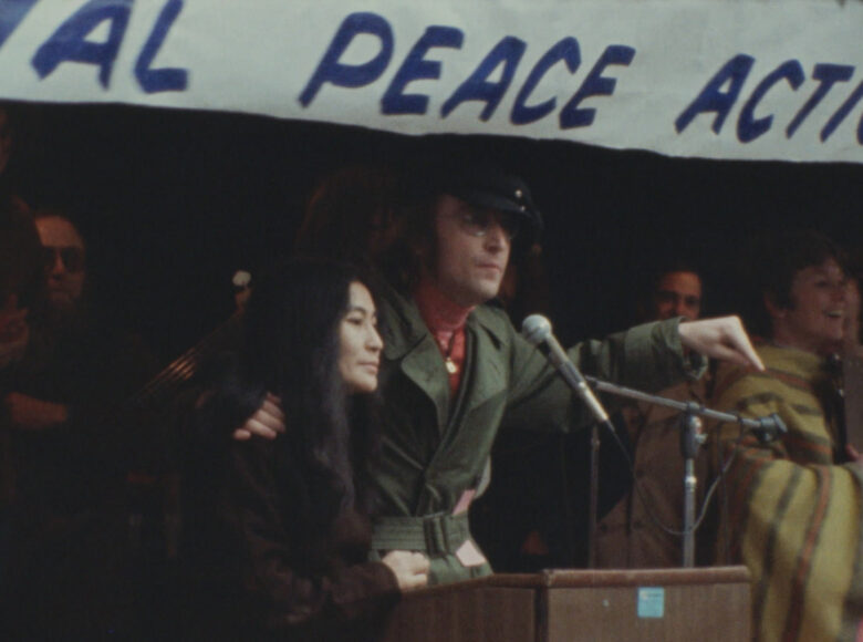 Yoko Ono i John Lennon stoją na trybunie. Nad nimi baner z częściowo widocznym hasłem na temat pokoju, napisanym po angielsku.
