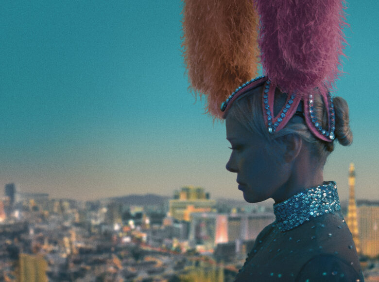 Na pierwszym planie zbliżenie na twarz i ramiona kobiety widziane z profilu. Kobieta ma bluzkę z cekinami, a na głowie przybranie z piór i cekinów. W tle widok na panoramę miasta i na wieczorne niebo w intensywnym niebieskim kolorze.