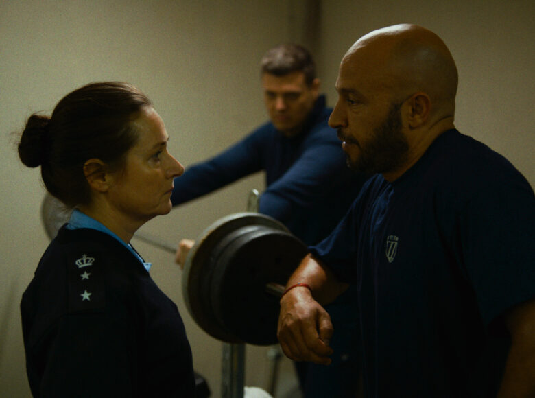 Kadr z filmu. Wnętrze siłowni. naprzeciwko siebie stoją kobieta i mężczyzna. Kobieta ma na sobie policyjny mundur, mężczyzna ma granatową koszulkę. W tle przy przyrządzie do ćwiczeń stoi drugi mężczyzna w granatowej bluzie.