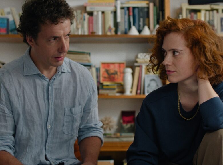 W pomieszczeniu młoda kobieta i młody mężczyzna siedzą obok siebie rozmawiając. Za nimi ściana z półkami na książki.