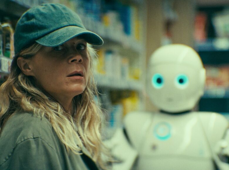 Wnętrze sklepu z kolorowymi towarami na półkach. Na pierwszym planie młoda dziewczyna w koszuli i czapce bejsbolówce. Za nią stoi robot.