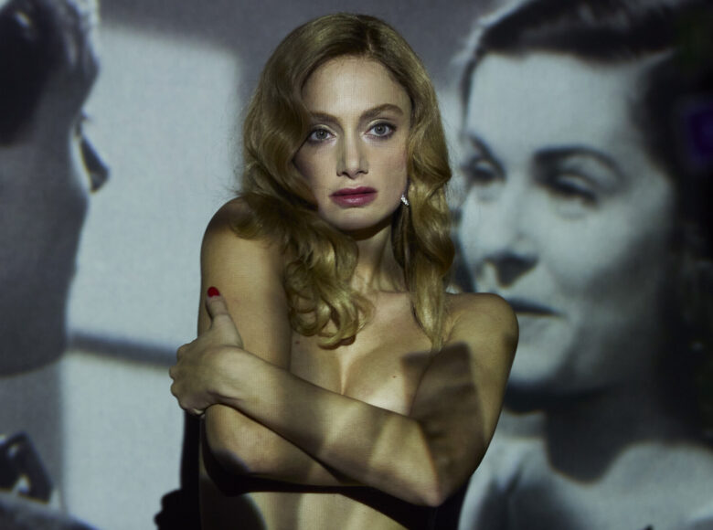 Młoda kobieta w ujęciu od pasa w górę. Ma długie włosy blond, makijaż i czerwone paznokcie oraz nagi tors. Piersi zasłania rękami. Za jej plecami ekran kinowy, na którym wyświetlany jest film.