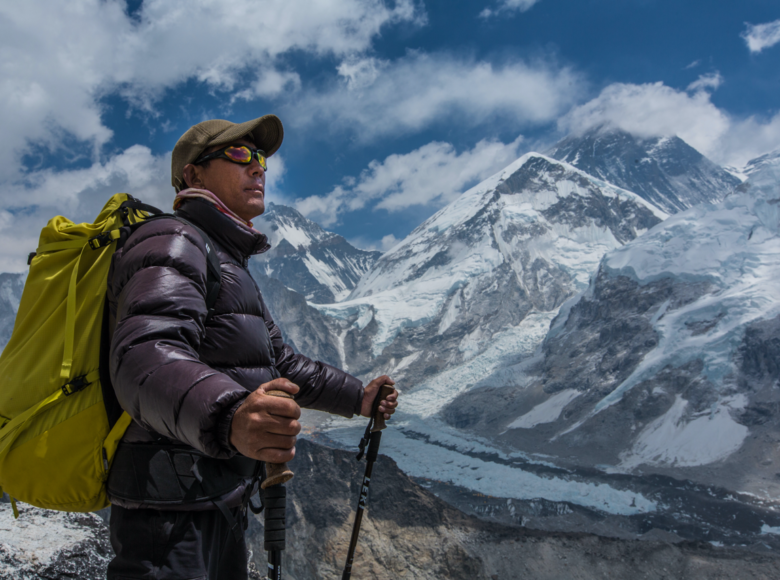 W tle szczyty Himalajów. Na pierwszym planie azjatycki mężczyzna w ortalionowej kurtce, z plecakiem na plecach i kijkami narciarskimi w dłoniach.