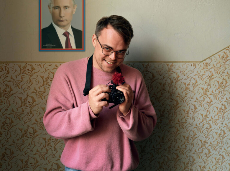 Młody, chytrze uśmiechnięty mężczyzna stoi w pomieszczeniu pod ścianą. W dłoniach trzyma aparat fotograficzny. Na ścianie wisi portret Putina.