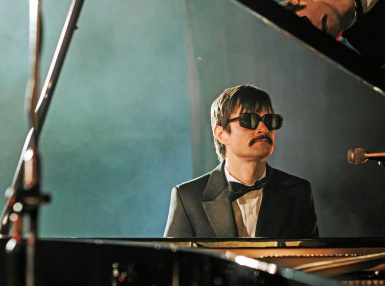 Sala koncertowa. Zbliżenie na mężczyznę grającego na fortepianie. Mężczyzna ma wąsy i okulary przeciwsłoneczne. Jest ubrany w czarny garnitur i czarną muszkę oraz białą koszulę.
