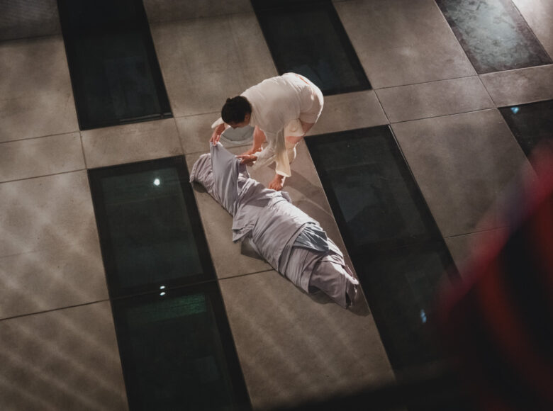 Spektakl tańca współczesnego. Na scenie tancerka owijaja wieloma warstwami tkaniny podłużny kształt leżący na podłodze. Kształt może kojarzyć się z mumią lub z ciałem człowieka w całunie.