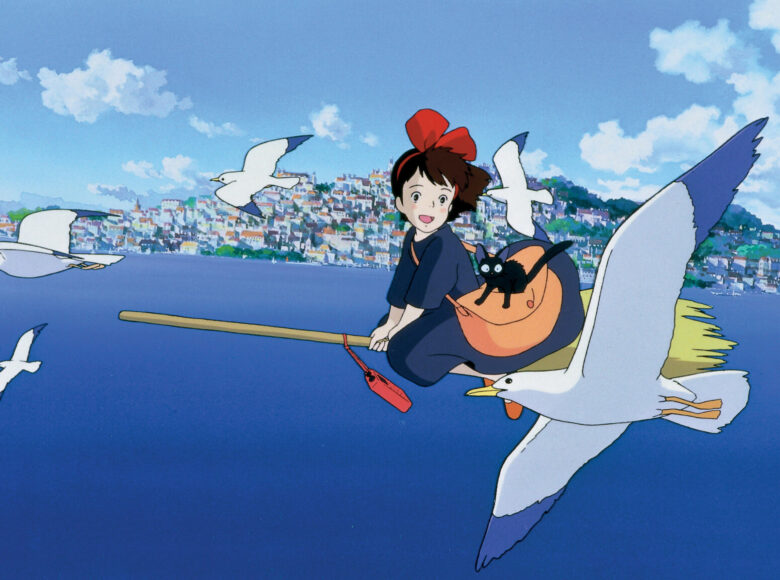 Kadr z filmu animowanego. Dziewczynka leci na miotle. Na ramieniu ma torbę, w której siedzi kot. Wokół latają ptaki. W dole widać morze, a na jego brzegu miasto.