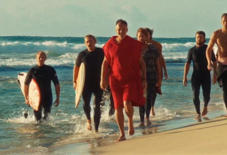 Kadr z filmu. Plaża. Popołudniowe światło słońca. Brzegiem morza idzie grupa osób - kilku mężczyzn i dwie kobiety. Niektórzy niosą deski surfingowe.