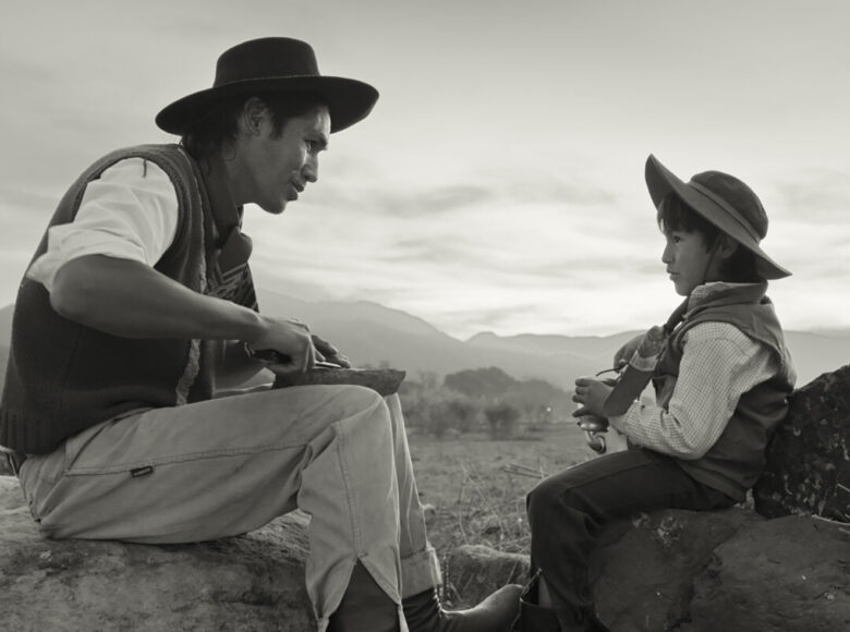 Kadr z filmu czarno białego. Mężczyzna i mały chłopiec siedzą naprzeciwko siebie na głazach, jedząc i rozmawiając. Za nimi widać pastwiska i wzgórza.