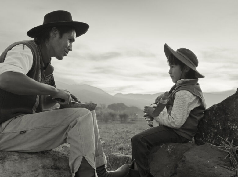 Kadr z filmu czarno białego. Mężczyzna i mały chłopiec siedzą naprzeciwko siebie na głazach, jedząc i rozmawiając. Za nimi widać pastwiska i wzgórza.