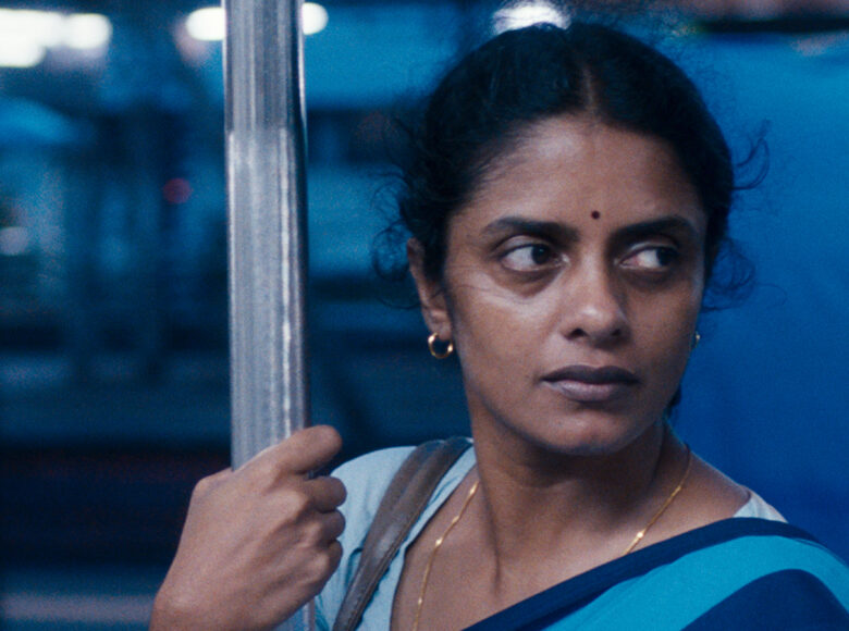 Kadr z filmu. Zbliżenie na twarz i ramiona hinduskiej kobiety. Kobieta przytrzymuje się poręczy autobusowej. Za nią w ciemnościach niedookreślone kształty i kilka punktów światła.