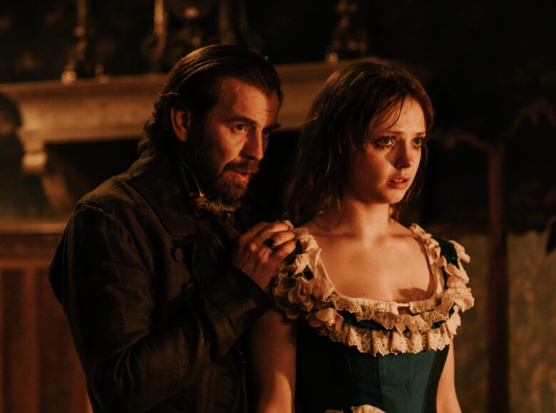 Kadr z filmu kostiumowego. dziewczyna i mężczyzna w starodawnych strojach stoją blisko siebie. Mężczyzna stoi nieco za dziewczyną, trzymając dłoń na jej ramieniu. Za nimi niewyraźne elementy architektury lub mebli wyłaniające się z ciemności.