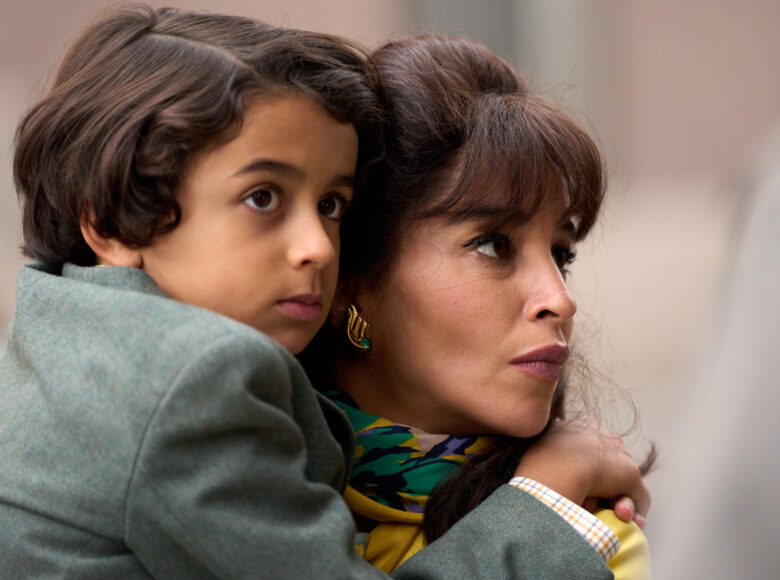 Zbliżenie na twarz i ramiona kobiety, trzymającej na rękach kilkuletniego chłopca. Kobieta ma elegancki makijaż i kolczyki, chłopiec nosi marynarkę.