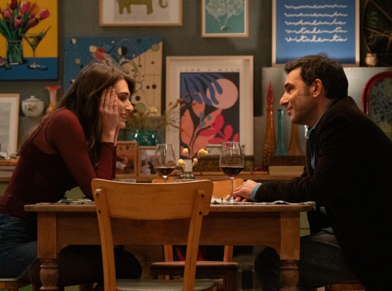 Kobieta i mężczyzna siedzą przy stole i piją czerwone wino, uśmiechając się do siebie. W tle za nimi ściana zawieszona wieloma kolorowymi obrazkami.