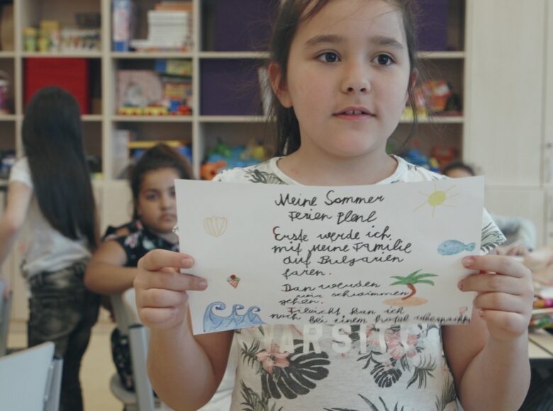 Klasa szkolna. Na pierwszym planie dziewczynka, trzymająca przed sobą kartkę pokrytą napisami i rysunkami. W tle kilkoro innych dzieci pracujących przy stolikach.