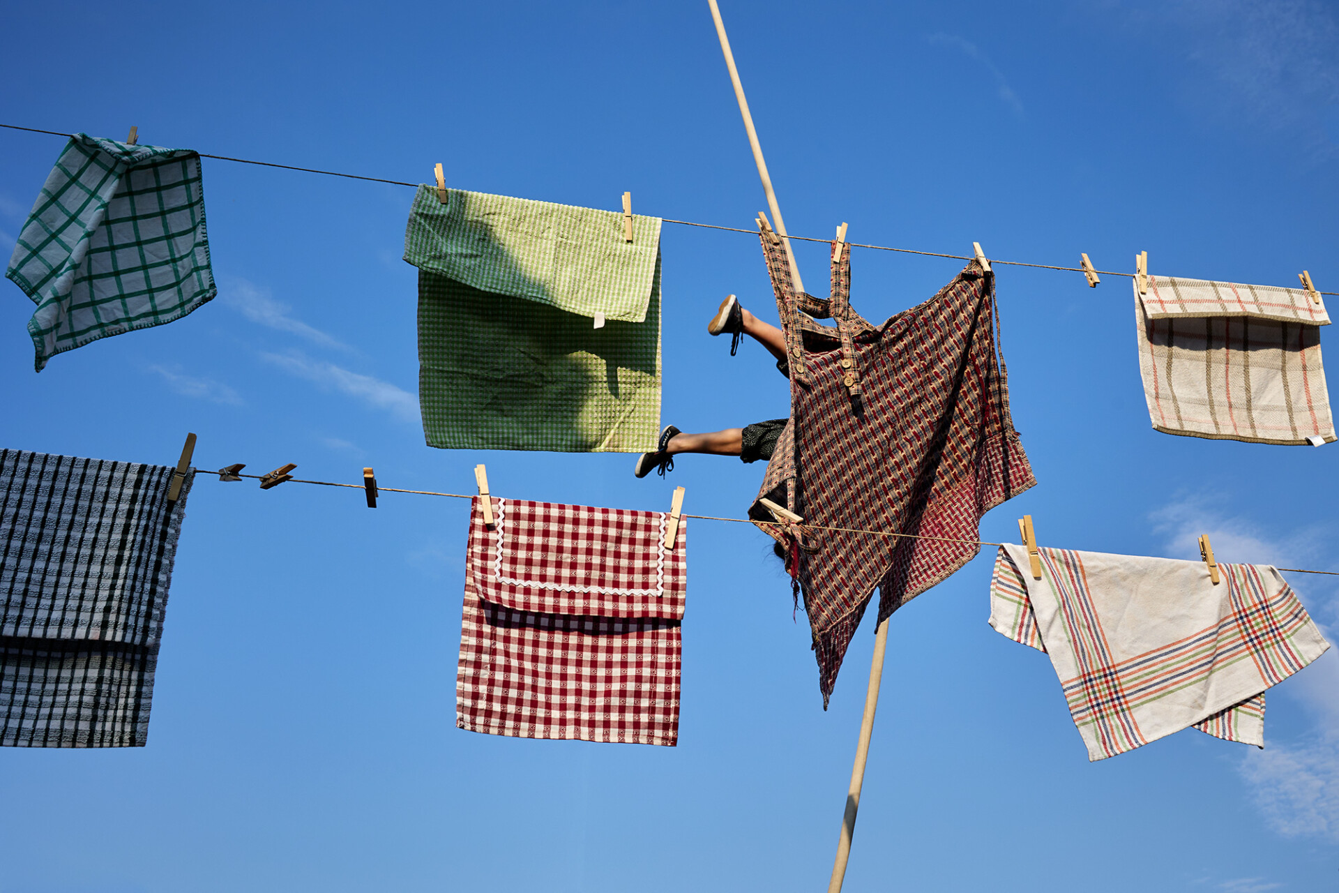 Kolorowa fotografia, o surrealistycznym charakterze, przedstawia wiszące na tle nieba pranie, poprzypinane do sznurków drewnianymi klamerkami. Zza jednej, przypiętej tymi klamerkami tkaniny, przypominającej ręcznik wystają nieproporcjonalnie małe (w stosunku do rozmiarów tego ręcznika) nogi człowieka - w sportowych butach z białą podeszwą i spodniach do kolan.