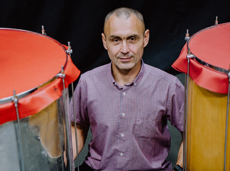 Między dwoma bębnami siedzi mężczyzna. Patrzy prosto w obiektyw. Za nim czarne tło.
