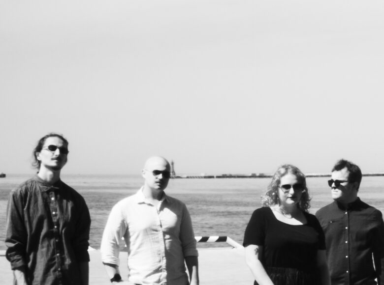 Zdjęcie czarno białe. Kobieta i trzech mężczyzn stoją obok siebie, pozując do zdjęcia. Mają na sobie lekkie ubrania i okulary słoneczne. Za nimi widać morze i niebo.