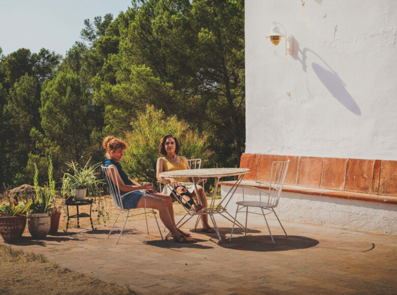 Lato. Na tarasie przed domem przy stole siedzą dwie kobiety. Za nimi drzewa i fragment akwenu.