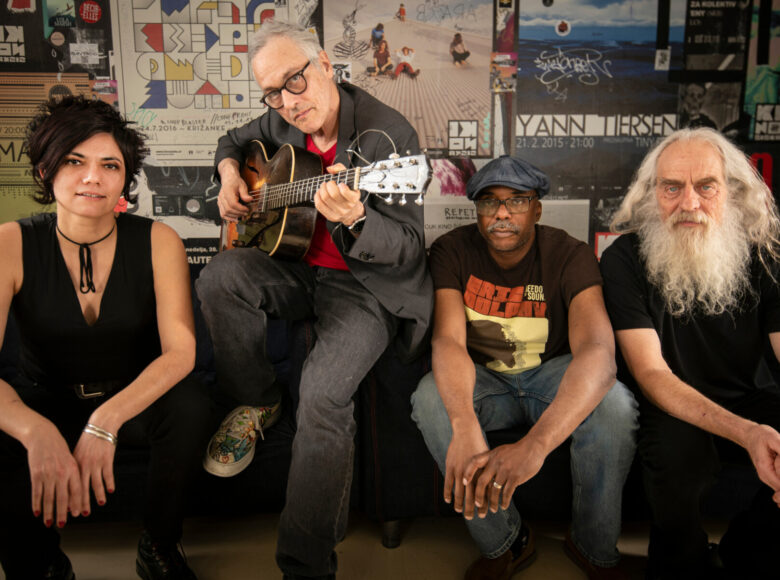 Na sofie. pozując do zdjęcia, siedzi młoda kobieta i trzech starszych mężczyzn. Jeden z nich trzyma gitarę. Za nimi ściana, na której wiszą plakaty do wydarzeń kulturalnych.