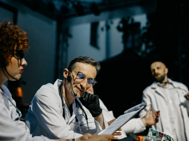 Przedstawienie teatralne. Na scenie przy stole siedzi grupa aktorów w białych kitlach lekarskich.