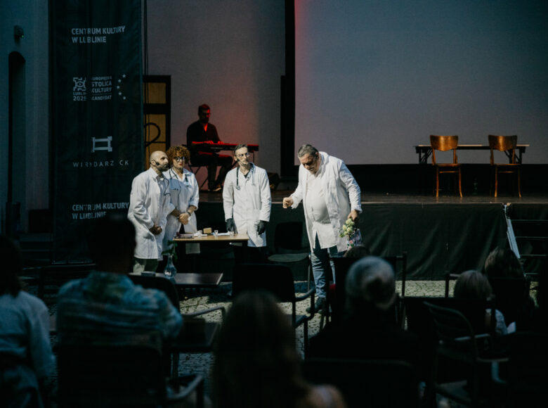 Przedstawienie teatralne. Na scenie stoi aktorka i trzech aktorów, wszyscy w białych kitlach lekarskich.