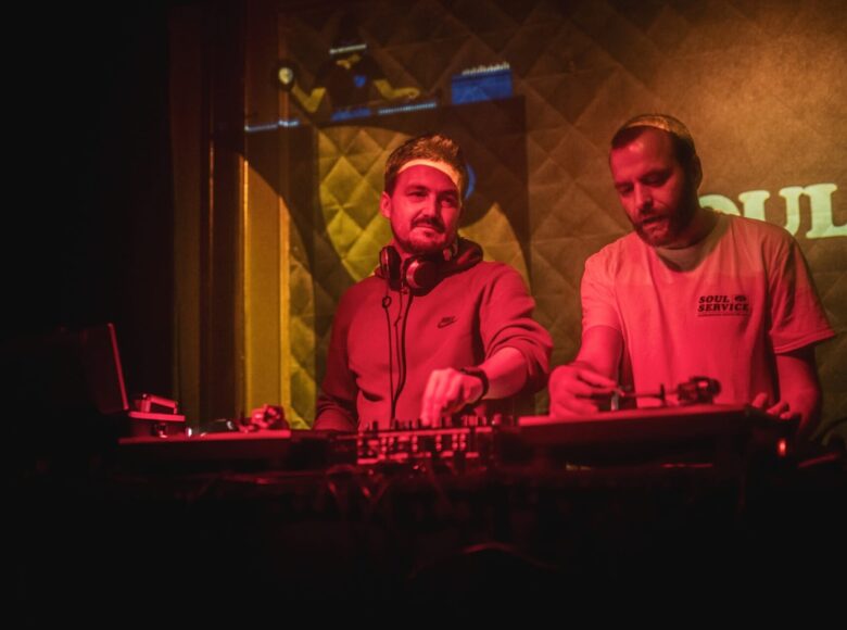 Koncert muzyki klubowej. Za stołem mikserskim stoi dwóch mężczyzn, puszczających muzykę.