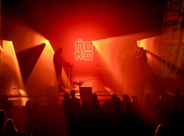 Koncert muzyki współczesnej, na scenie kilku muzyków. Scena jest intensywnie oświetlona czerwonym i żółtym światłem reflektorów.