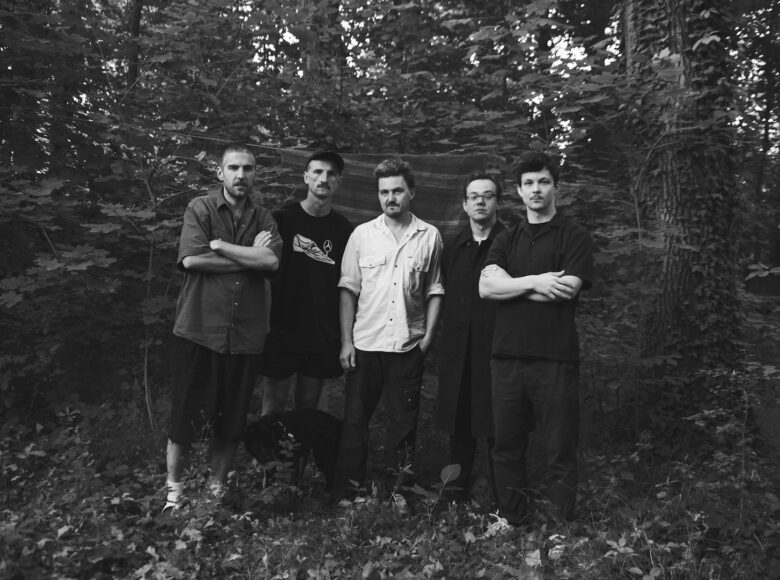 Fotografia czarno biała. Lato. W lesie lub parku wśród drzew stoi pięciu mężczyzn, pozując do fotografii.