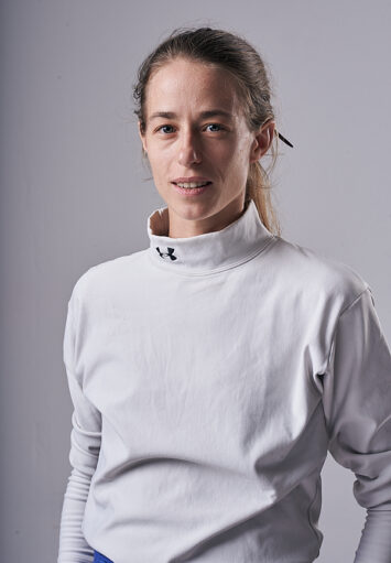 Fotograficzny portret młodej kobiety na neutralnym, szarym tle. Kobieta ma długie, jasne włosy związane z tyłu. Ubrana jest w białą bluzę.