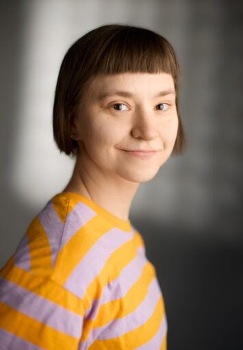 Fotograficzny portret młodej kobiety na neutralnym, szarym tle. Kobieta ma krótkie, ciemne włosy, brązowe oczy, ubrana jest w bluzkę w żółto-szare paski.