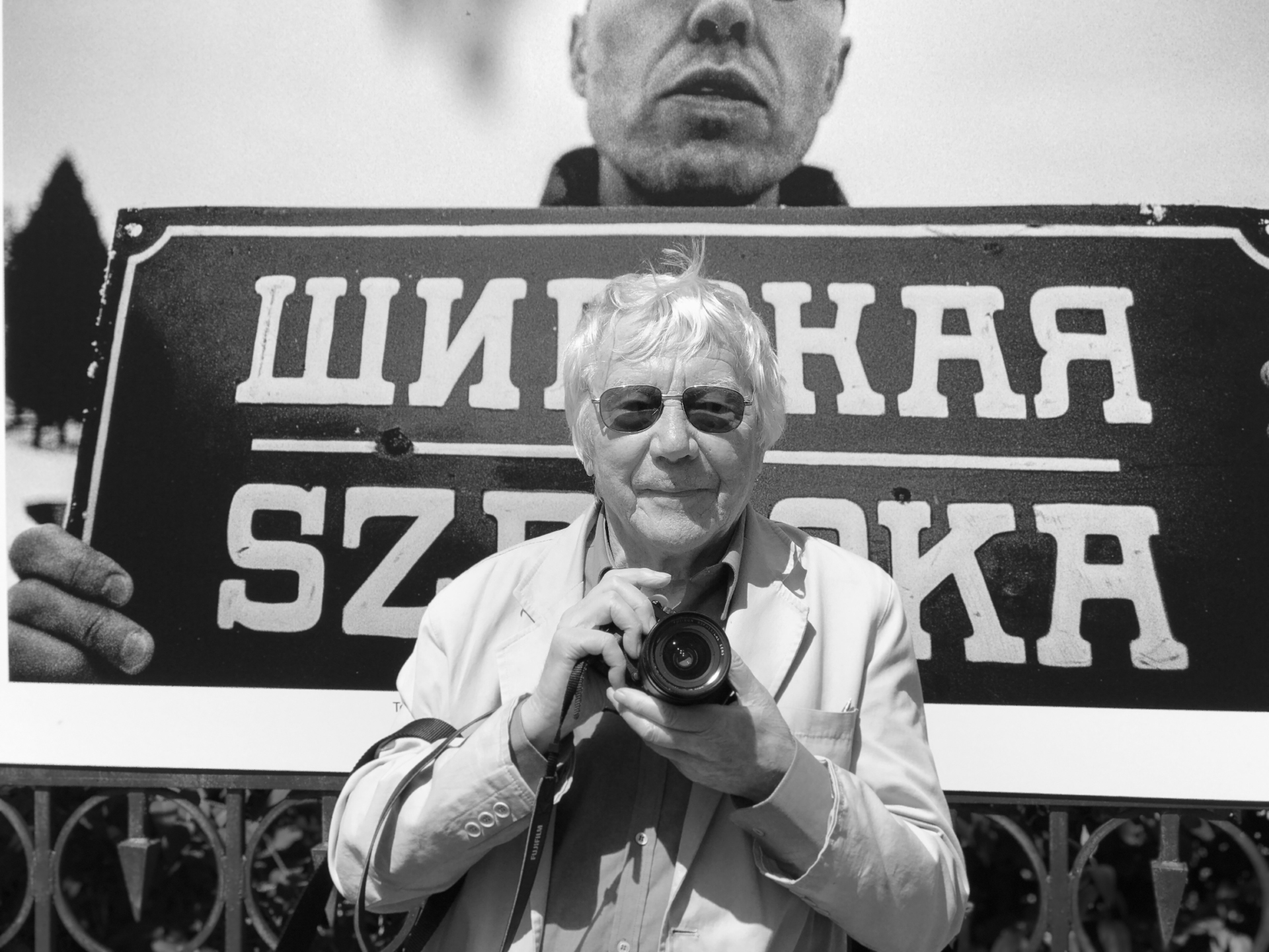 Zdjęcie czarno białe. Przedstawia fotografika Tadeusza Rolke z aparatem fotograficznym w dłoniach. W tle za Rolkem dużowormatowa fotografia przedstawiająca Tomasza Pietrasiewicza, dyrektora Teatru NN trzymającego w ręku historyczną tablicę z nazwą ulicy Szeroka pisaną po polsku i cyrylicą.