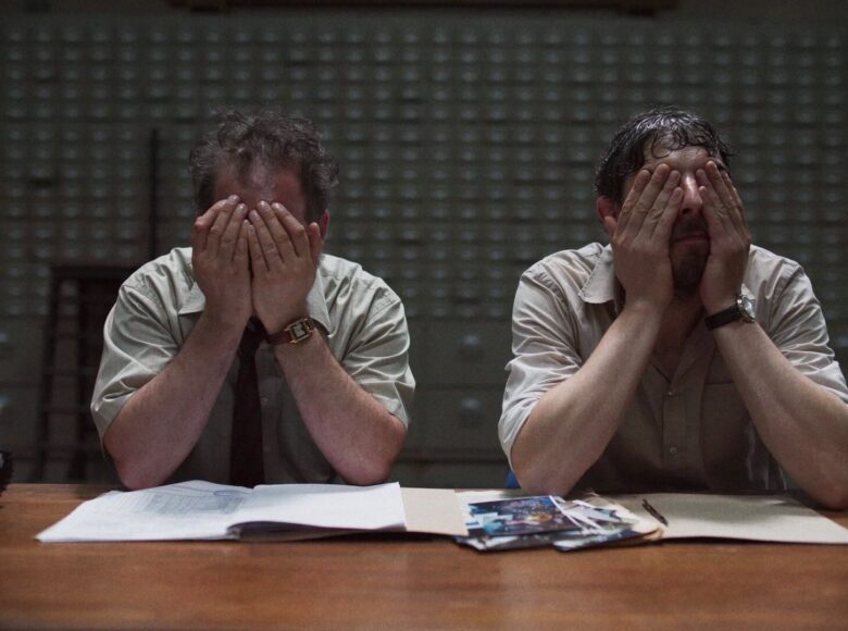 Kadr z filmu. W pomieszczeniu biurowym przy biurku siedzą obok siebie dwaj mężczyźni w identycznej pozie. Każdy opiera łokcie na blacie i zakrywa swoją twarz dłońmi.