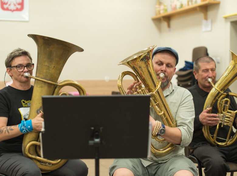 W pomieszczeniu trzech mężczyzn grających na dużych instrumentach dętych, jak tuby czy waltornie.