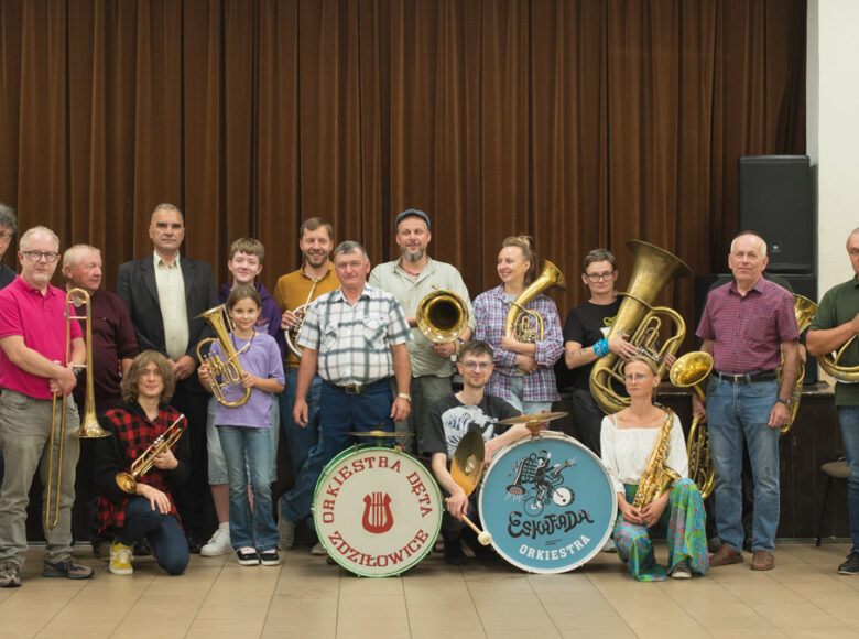 Na scenie do fotografii pozuje grupa osób płci obojga i w różnym wieku, z instrumentami dętymi i z bębnami.