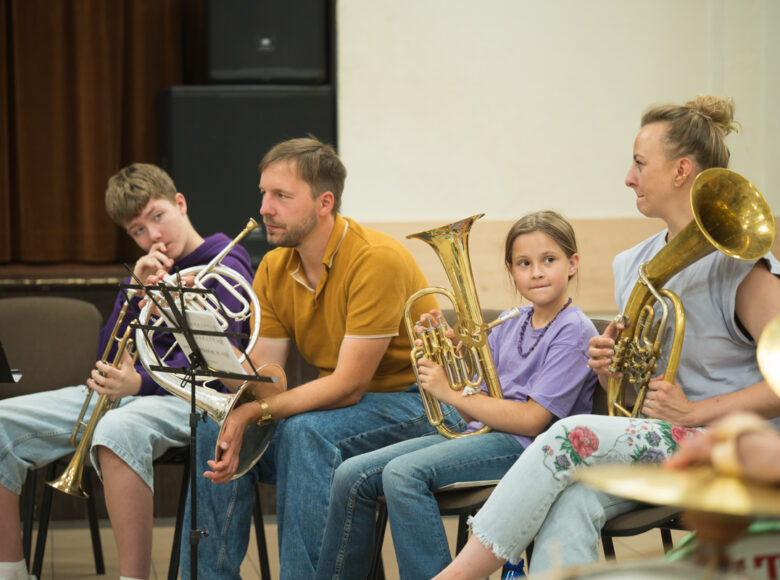W pomieszczeniu w rzędzie obok siebie siedzą kobieta, mężczyzna, dziewczynka i chłopiec. Wszyscy trzymają w rękach instrumenty dęte.