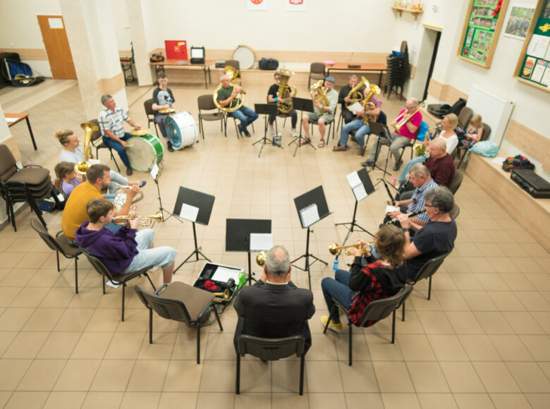 W dużej sali grupa osób siedzi w kręgu na krzesłach. Grają na instrumentach dętych i na bębnach.
