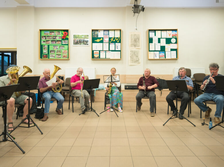 W pomieszczeniu grupa osób grających na różnych instrumentach dętych.