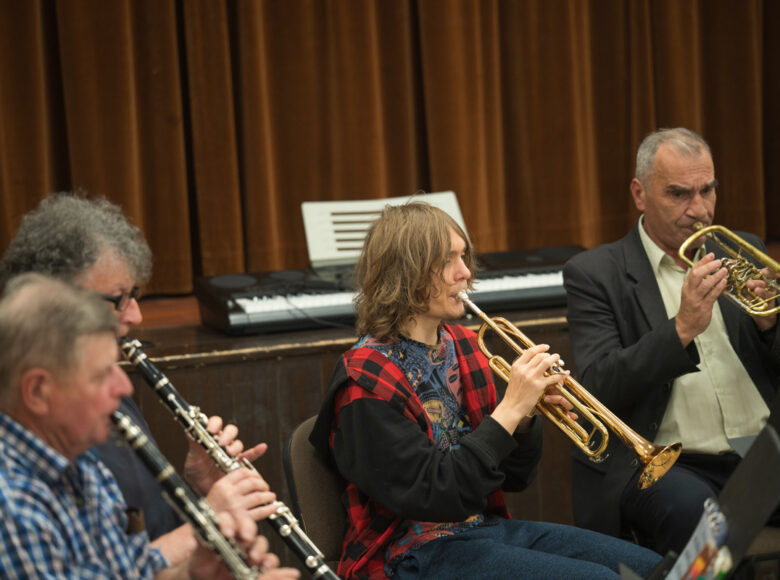 W pomieszczeniu trzech mężczyzn i chłopiec, siedzący obok siebie. Grają na puzonach i klarnetach.