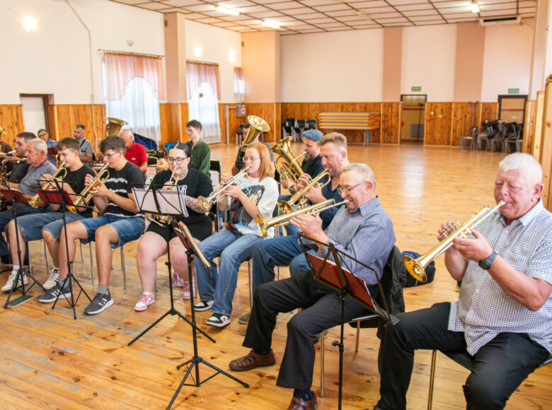W sali w rzędzie na krzesłach siedzi grupa osób, grających na instrumentach dętych.