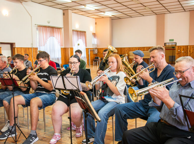 W sali w rzędzie na krzesłach siedzi grupa osób, grających na instrumentach dętych.