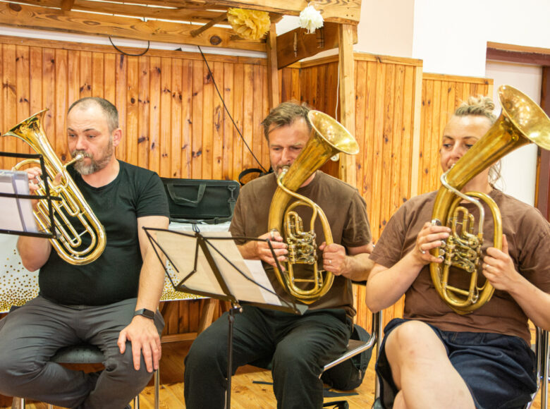 W pomieszczeniu trzy osoby grające na trąbkach, siedzące obok siebie. Za nimi widać ścianę pokrytą boazerią.