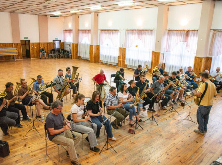 Zdjęcie z lotu ptaka. W sali na krzesłach w rzędach siedzi duża grupa osób, grających na instrumentach dętych.