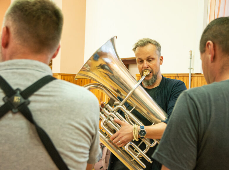 W pomieszczeniu mężczyzna grający na tubie. Przed nim stoją dwaj inni mężczyźni.