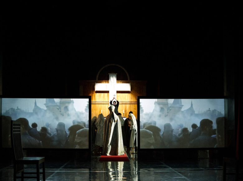 Spektakl teatralny. W scenografii przedstawiającej wnętrze kościoła grupa osób płci obojga ubranych w habity i sutanny, odgrywających mszę. Obok nich stoją dwa ekrany, na których wyświetlany jest widok na tłum ludzi przed budynkiem kościoła.