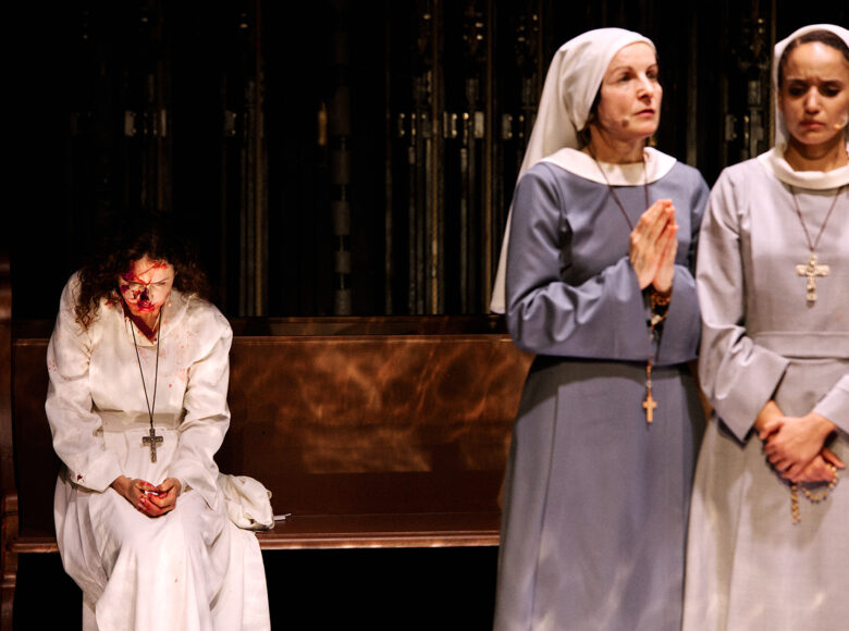 Przedstawienie teatralne. Scenografia przedstawia wnętrze kościoła. Na pierwszym panie stoją obok siebie dwie kobiety w szarych habitach. Jedna z nich trzyma dłonie w geście modlitwy. Za nimi na ławce siedzi kobieta w białym habicie. Na twarzy i rękach ma rozmazane plamy czerwonej cieczy, kojarzące się z krwią.